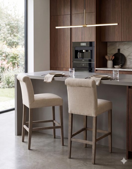 Banqueta alta de diseño contemporáneo con estructura de madera maciza en acabado natural. Presenta un asiento y respaldo integrado, tapizado en pana color claro con textura sutil. Incluye apoyapiés perimetral de madera para mayor estabilidad y confort