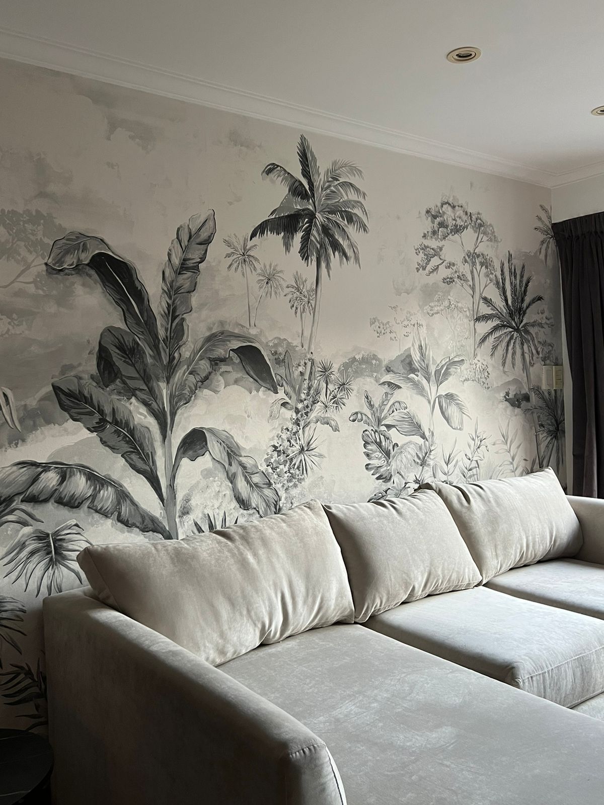 Sofá esquinero tapizado en tela beige claro, con tres cojines del mismo tono, ubicado frente a una pared decorada con un mural de estilo tropical en blanco y negro que presenta hojas y palmeras de gran tamaño