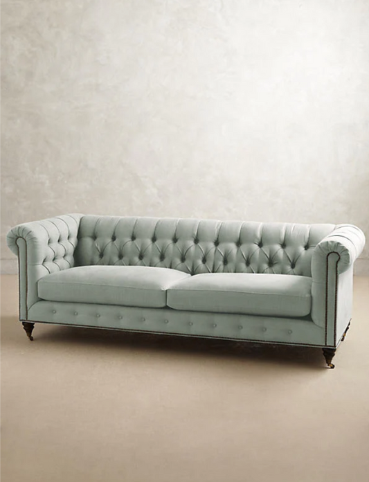 Sofá clásico Chesterfield, tapizado en pana, con respaldo y apoyabrazos capitoné, líneas elegantes y sofisticadas, patas de madera oscura tallada, sobre fondo blanco, ideal para decorar livings, oficinas o salas de estilo tradicional con un toque de lujo.