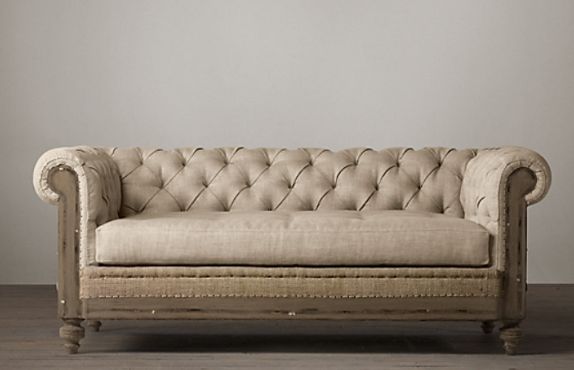 Fotografía editorial de interiores que muestra un living sofisticado y tradicional. El protagonista es un sillón Chesterfield de tres cuerpos, tapizado en lino texturizado color beige, con capitoné profundo, brazos enrollados y una estructura de madera envejecida con detalles de tachuelas. El sillón se apoya sobre una alfombra persa antigua sobre piso de madera. Al fondo, una gran biblioteca empotrada de piso a techo, hecha de madera oscura, está llena de libros y objetos decorativos como jarrones de latón