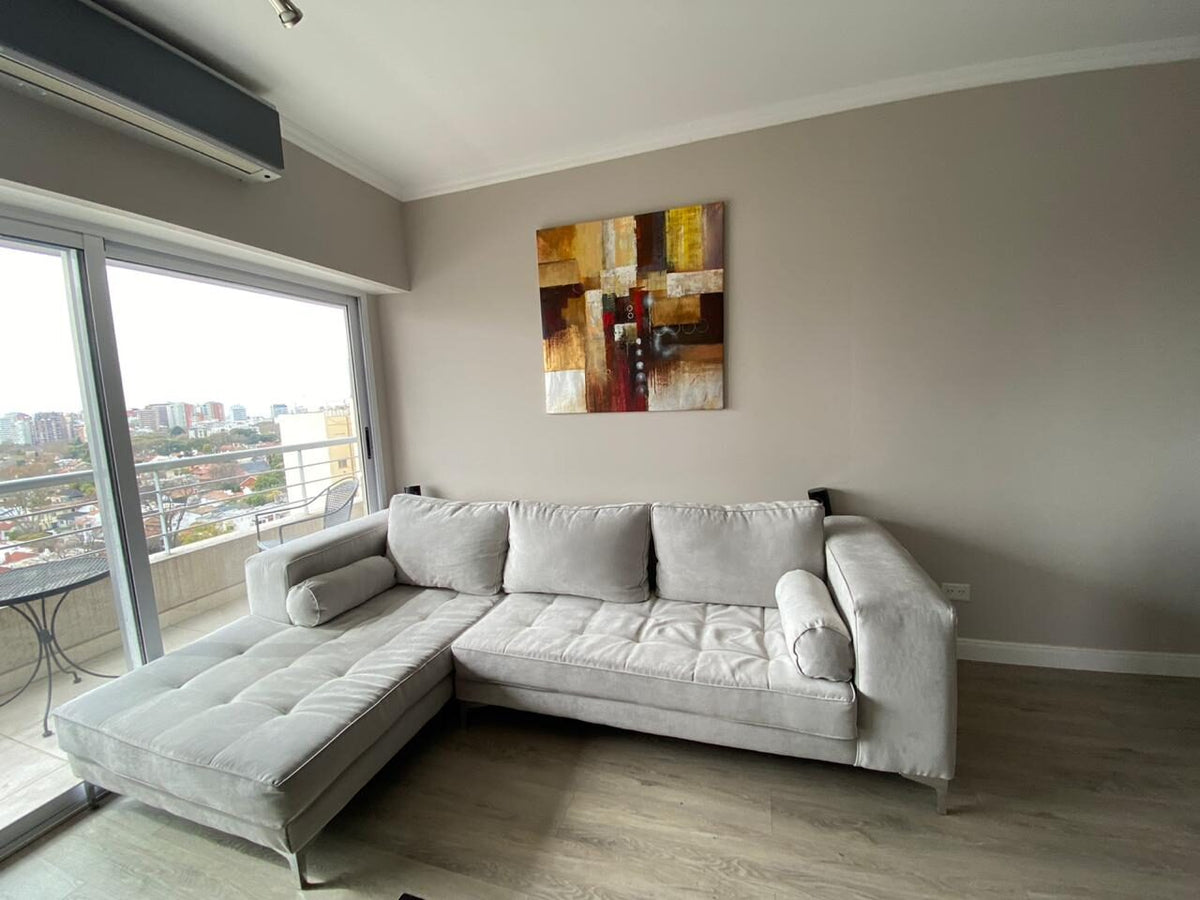 Esquinero Bambú gris perla con patas cromadas y chaise, frente al ventanal del departamento de Alejandra con vista a la ciudad