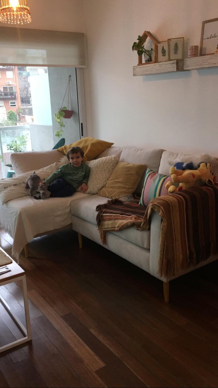 Sillón Esquinero Bélgica beige con patas de madera en el living con balcón de Antonella, con almohadones mostaza y una manta tejida