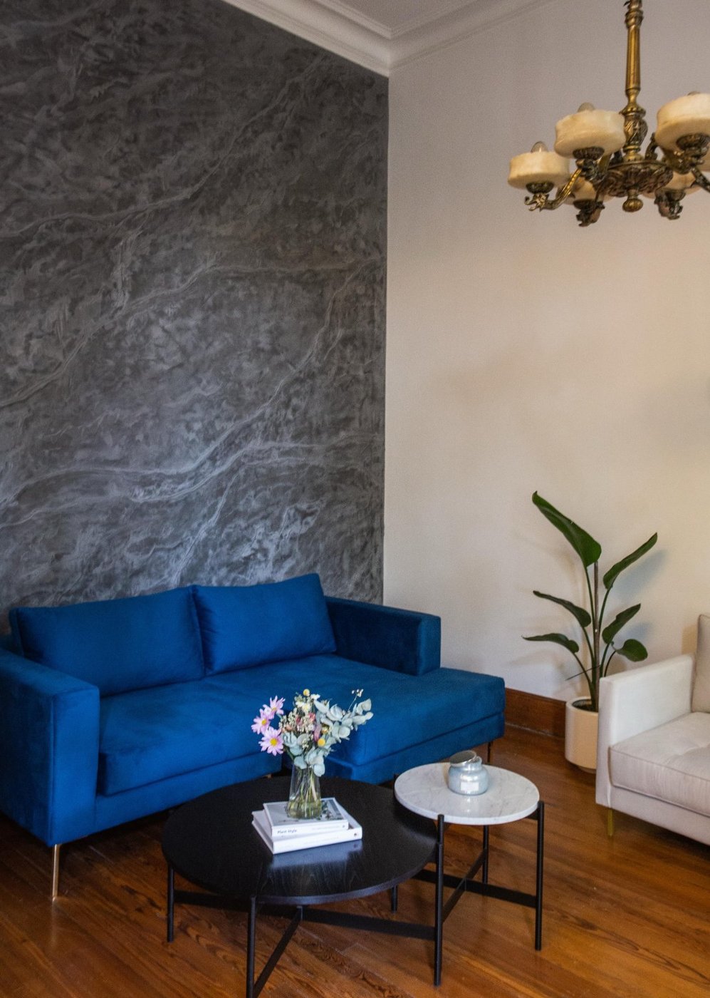 Esquinero Magnolia azul con chaise y patas doradas frente a una pared de textura gris, con araña antigua de bronce y piso de madera