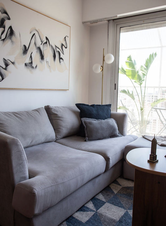 Sillón esquinero tapizado en tono gris claro, con almohadones decorativos en azul y gris, acompañado de una mesa auxiliar de madera sobre una alfombra geométrica en tonos azules y blancos, en un living luminoso y moderno