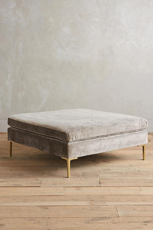 Banqueta rectangular tapizada en con patas doradas. Su diseño compacto y moderno la vuelve ideal para espacios reducidos o como apoya pies.