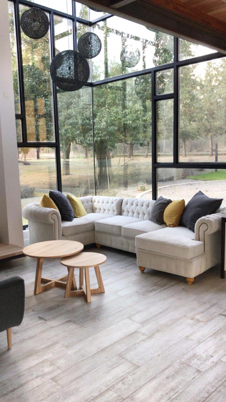 Un sillón esquinero amplio en color beige claro con diseño capitoné, decorado con almohadones grises y amarillos, ubicado en un living con ventanales de vidrio que permiten ver un entorno natural exterior.