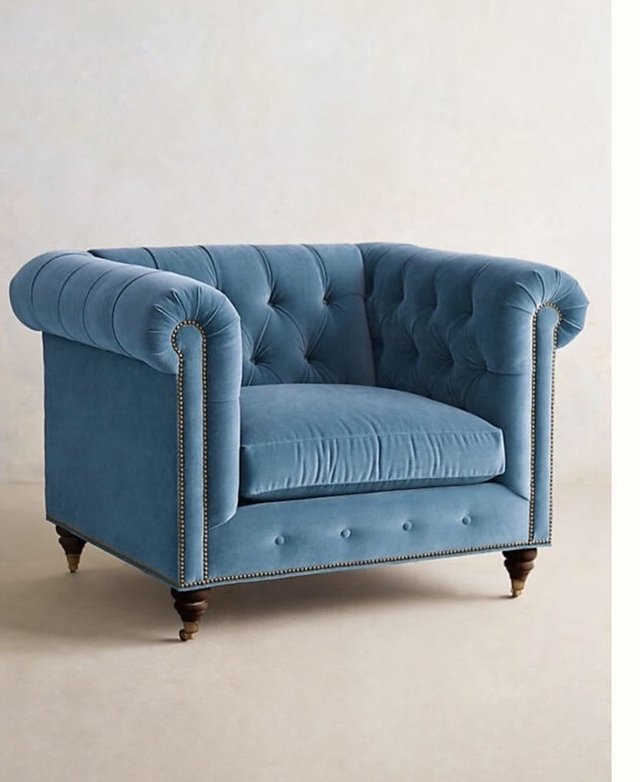 Sillón individual estilo Chesterfield tapizado en color celeste, con capitoné en respaldo y apoyabrazos, tachas decorativas y patas de madera torneadas.