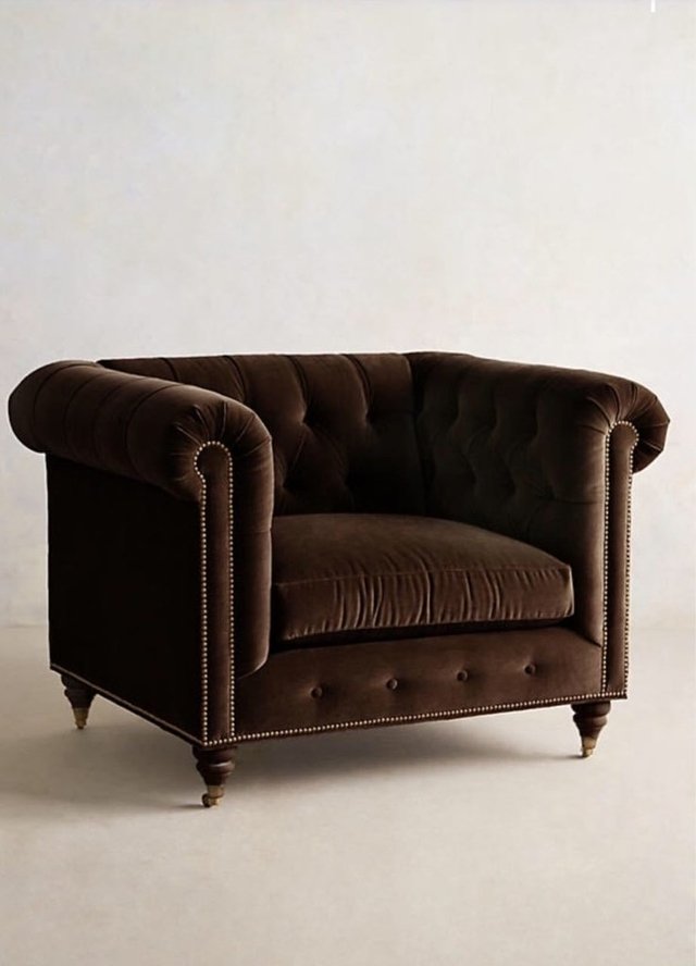 Sillón individual estilo Chesterfield tapizado en color marron, con capitoné en respaldo y apoyabrazos, tachas decorativas y patas de madera torneadas.