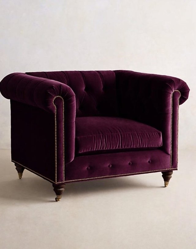 Sillón individual estilo Chesterfield tapizado en color violeta, con capitoné en respaldo y apoyabrazos, tachas decorativas y patas de madera torneadas.