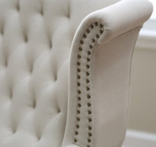 Sillón estilo clásico tapizado en color gris claro, con respaldo capitoné alto, orejas curvas, apoyabrazos redondeados y tachas decorativas, patas de madera talladas