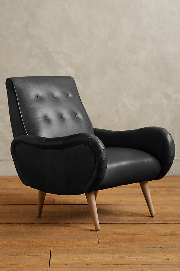 Sillón de un cuerpo estilo retro tapizado en negro, con respaldo bajo capitoné, apoyabrazos curvos y patas de madera cónicas.