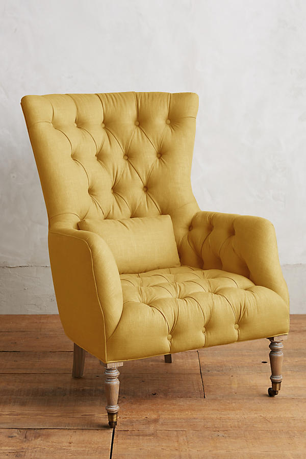 Sillón individual tapizado en amarillo, con respaldo alto y capitoné, cojín lumbar suelto, brazos curvos y patas delanteras torneadas en madera clara.
