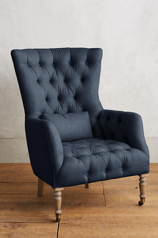 Sillón individual tapizado en azul, con respaldo alto y capitoné, cojín lumbar suelto, brazos curvos y patas delanteras torneadas en madera clara.