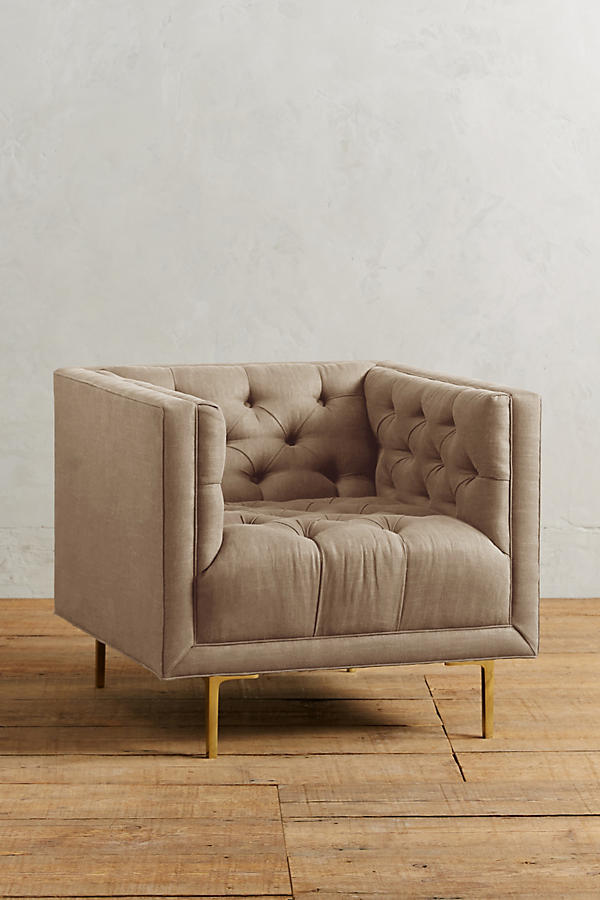 Sillón individual tapizado en beige, de forma cúbica con respaldo y brazos rectos, capitoné de botones en todo el interior y patas delgadas de madera.