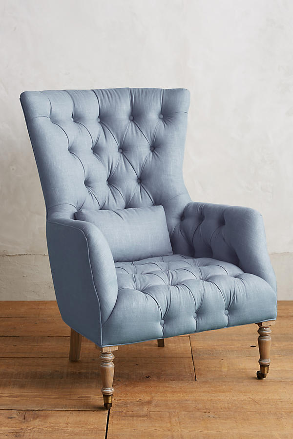 Sillón individual tapizado en celeste, con respaldo alto y capitoné, cojín lumbar suelto, brazos curvos y patas delanteras torneadas en madera clara.