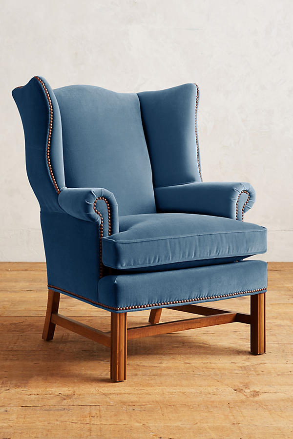 Sillón individual tapizado en terciopelo celeste, con respaldo alto y orejero, cojines sueltos en asiento y respaldo, y base de madera vista con patas cruzadas.