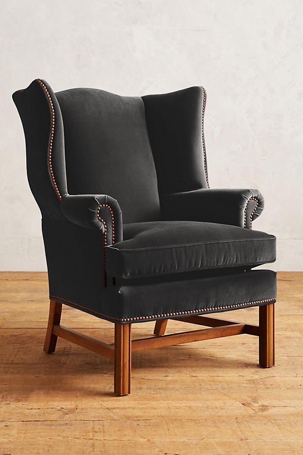 Sillón individual tapizado en terciopelo gris, con respaldo alto y orejero, cojines sueltos en asiento y respaldo, y base de madera vista con patas cruzadas.