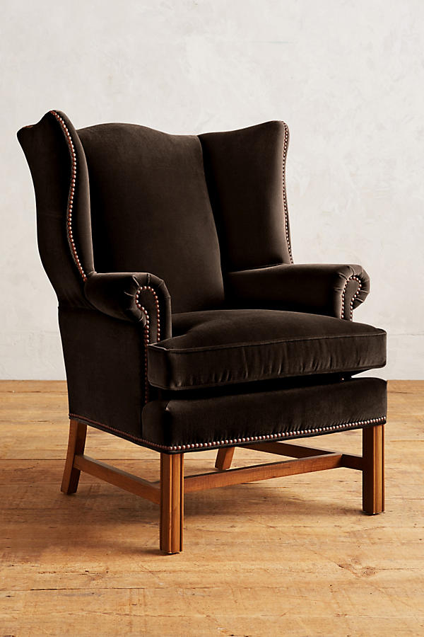 Sillón individual tapizado en terciopelo marron, con respaldo alto y orejero, cojines sueltos en asiento y respaldo, y base de madera vista con patas cruzadas.