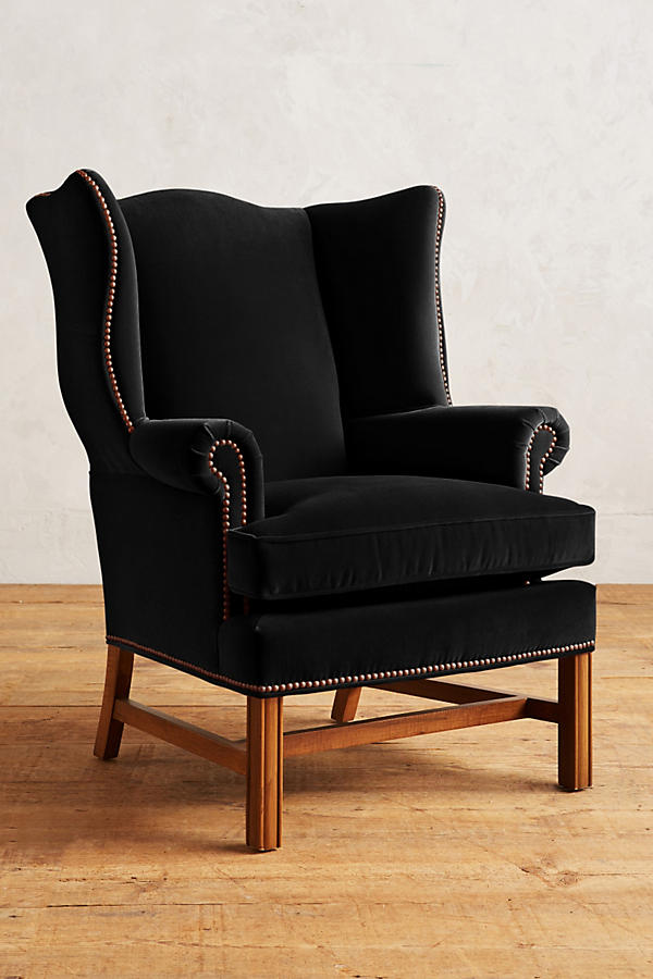 Sillón individual tapizado en terciopelo negro, con respaldo alto y orejero, cojines sueltos en asiento y respaldo, y base de madera vista con patas cruzadas.