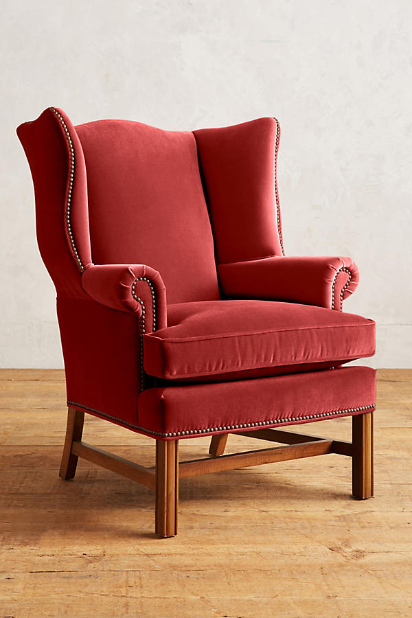 Sillón individual tapizado en terciopelo rojo, con respaldo alto y orejero, cojines sueltos en asiento y respaldo, y base de madera vista con patas cruzadas.