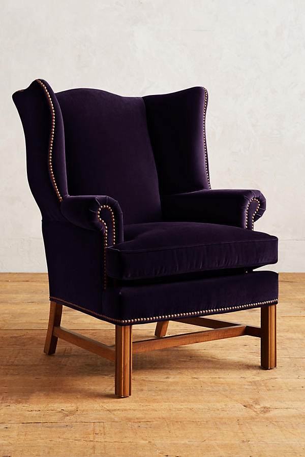 Sillón individual tapizado en terciopelo violeta, con respaldo alto y orejero, cojines sueltos en asiento y respaldo, y base de madera vista con patas cruzadas.