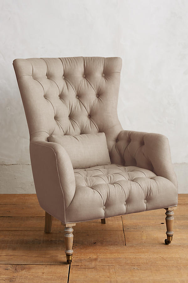 Sillón individual tapizado en beige, con respaldo alto y capitoné, cojín lumbar suelto, brazos curvos y patas delanteras torneadas en madera clara.