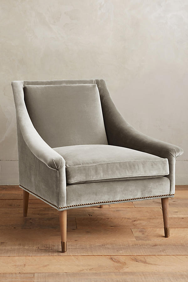 Sillón individual tapizado en gris, de líneas rectas con respaldo y asiento acolchados, brazos delgados y patas de madera rectas con detalle de tachas decorativas en la parte inferior.