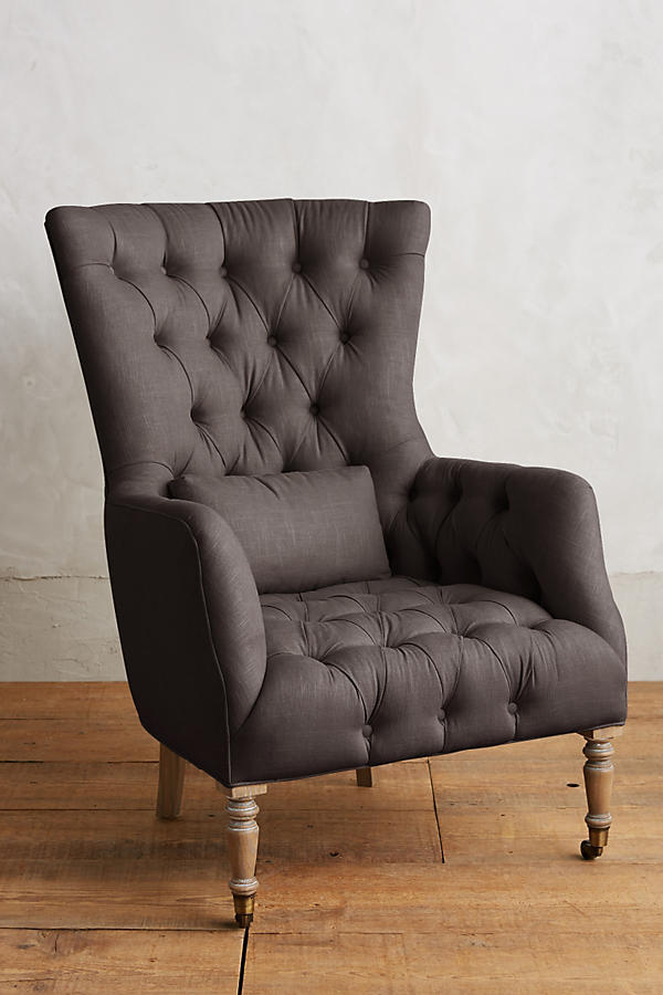 Sillón individual tapizado en gris, con respaldo alto y capitoné, cojín lumbar suelto, brazos curvos y patas delanteras torneadas en madera clara.