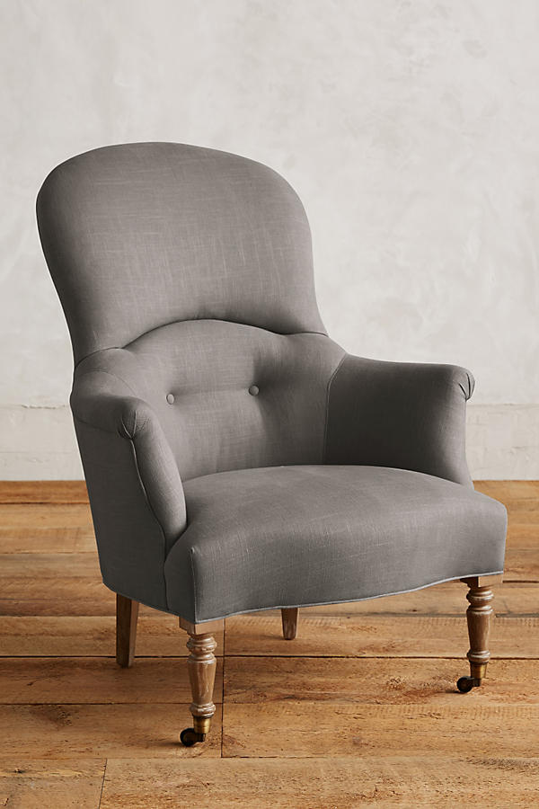 Sillón individual de diseño tradicional en tono gris claro, con respaldo curvo, apoyabrazos tallados y patas delanteras de madera con ruedas.