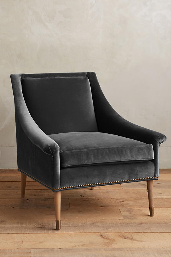 Sillón individual tapizado en gris claro, de líneas rectas con respaldo y asiento acolchados, brazos delgados y patas de madera rectas con detalle de tachas decorativas en la parte inferior.