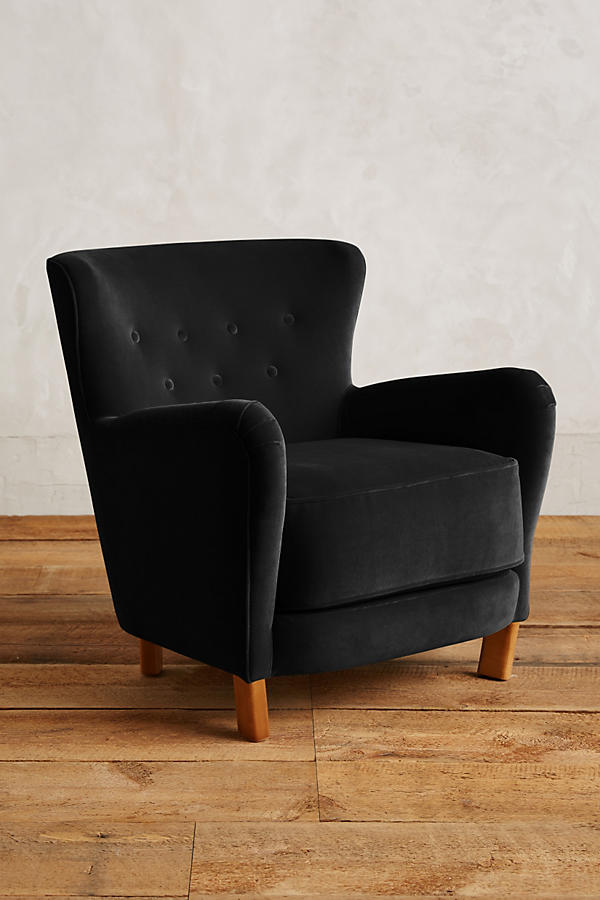 Sillón individual tapizado en negro, con respaldo suavemente inclinado y capitoné de botones, brazos redondeados y patas cortas de madera.