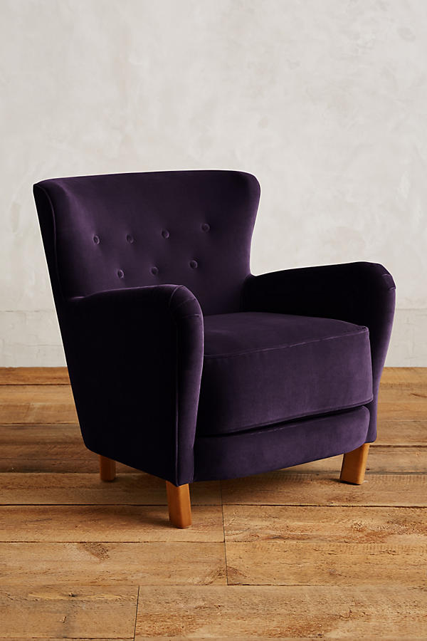 Sillón individual tapizado en violeta, con respaldo suavemente inclinado y capitoné de botones, brazos redondeados y patas cortas de madera.
