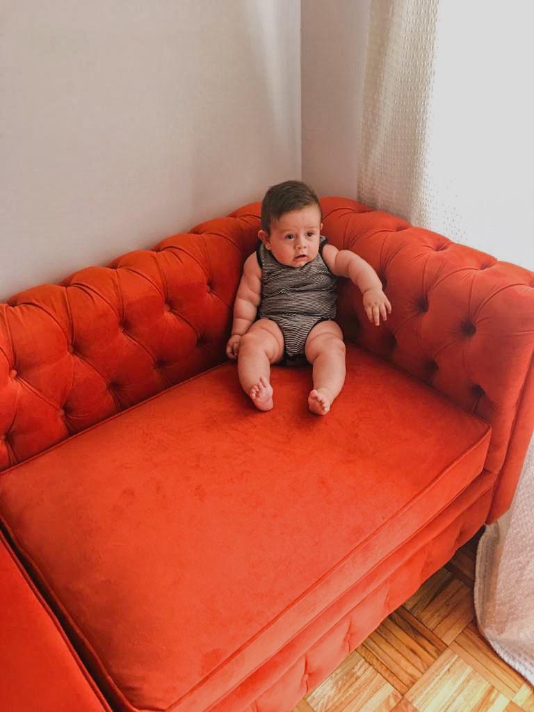 Un amplio sillón en forma de "L" tapizado en terciopelo rojo anaranjado ocupa gran parte de la imagen. El diseño del sillón es clásico, con respaldos y brazos acolchados de capitoné. En una de las esquinas del sillón, se encuentra un bebé pequeño, lo que acentúa la gran escala del mueble. La luz natural entra desde una ventana con cortina blanca al lado derecho, iluminando parte del sillón y del piso de madera.