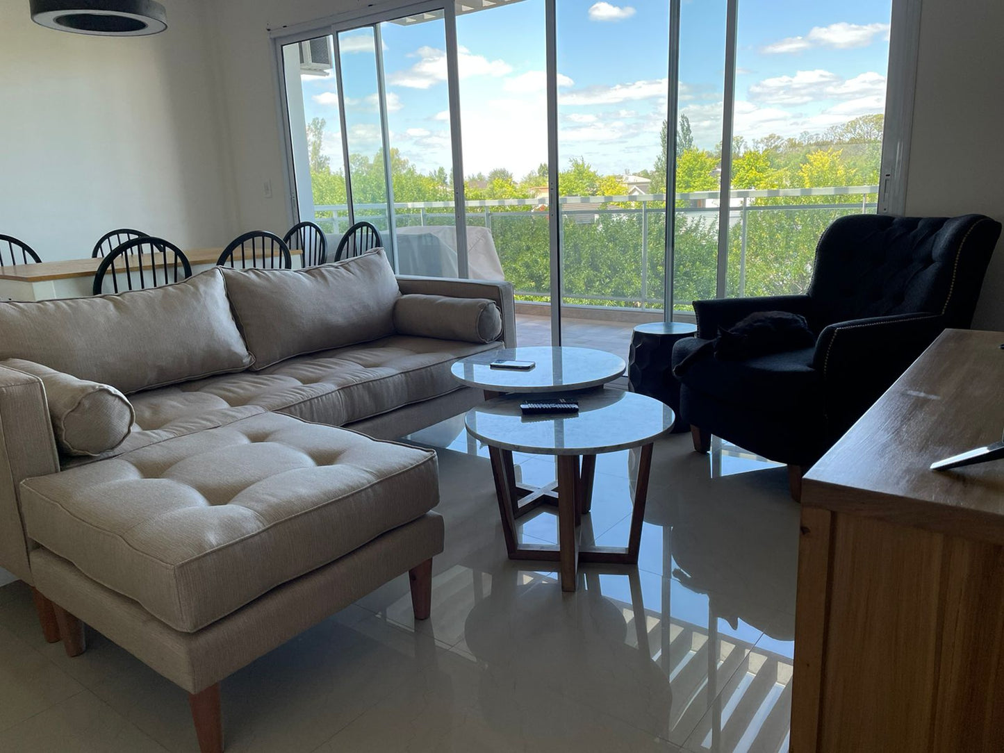 Sofá esquinero tapizado en tono beige claro con chaise longue y patas de madera, de diseño moderno y confortable, ideal para un living luminoso con amplios ventanales