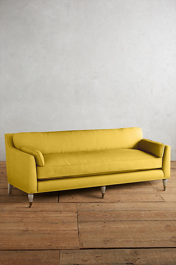 Fotografía de diseño de interiores estilo editorial que muestra un living moderno e industrial sofisticado. En el centro se destaca un sillón de tres cuerpos tapizado en lino gris claro con patas torneadas blancas y rueditas de bronce, idéntico al modelo de referencia. Está ubicado sobre una alfombra de diseño clásico envejecido superpuesta a una gran alfombra de yute