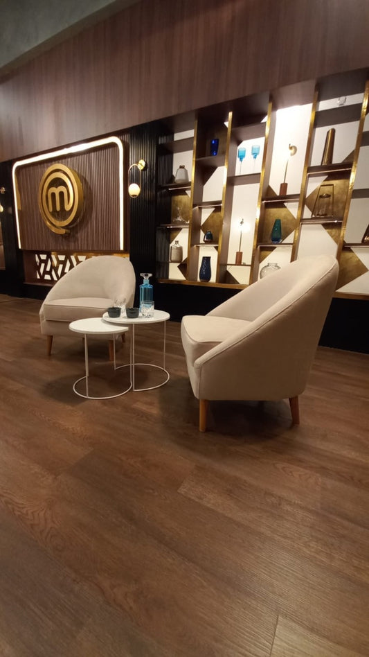 Dos sillones modernos de tapizado beige claro con patas de madera, dispuestos en un área de espera con suelo de madera oscura. Entre ellos hay dos mesas pequeñas redondas, una con una botella y vaso de agua. Al fondo se observa una pared decorativa con estanterías iluminadas y un gran logo dorado de MasterChef