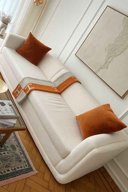 Sillón de tres cuerpos color blanco con dos almohadones de terciopelo terracota y una manta en tonos naranjas, ubicado en un living luminoso con mesa ratona de vidrio y alfombra persa.