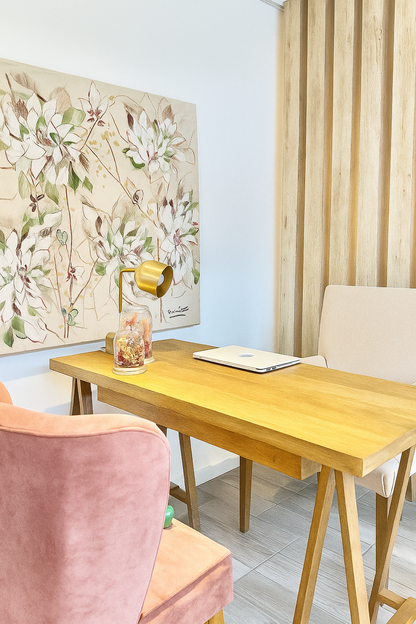 Escritorio de madera con silla beige y silla rosa, decorado con un cuadro floral en tonos claros colgado en la pared.