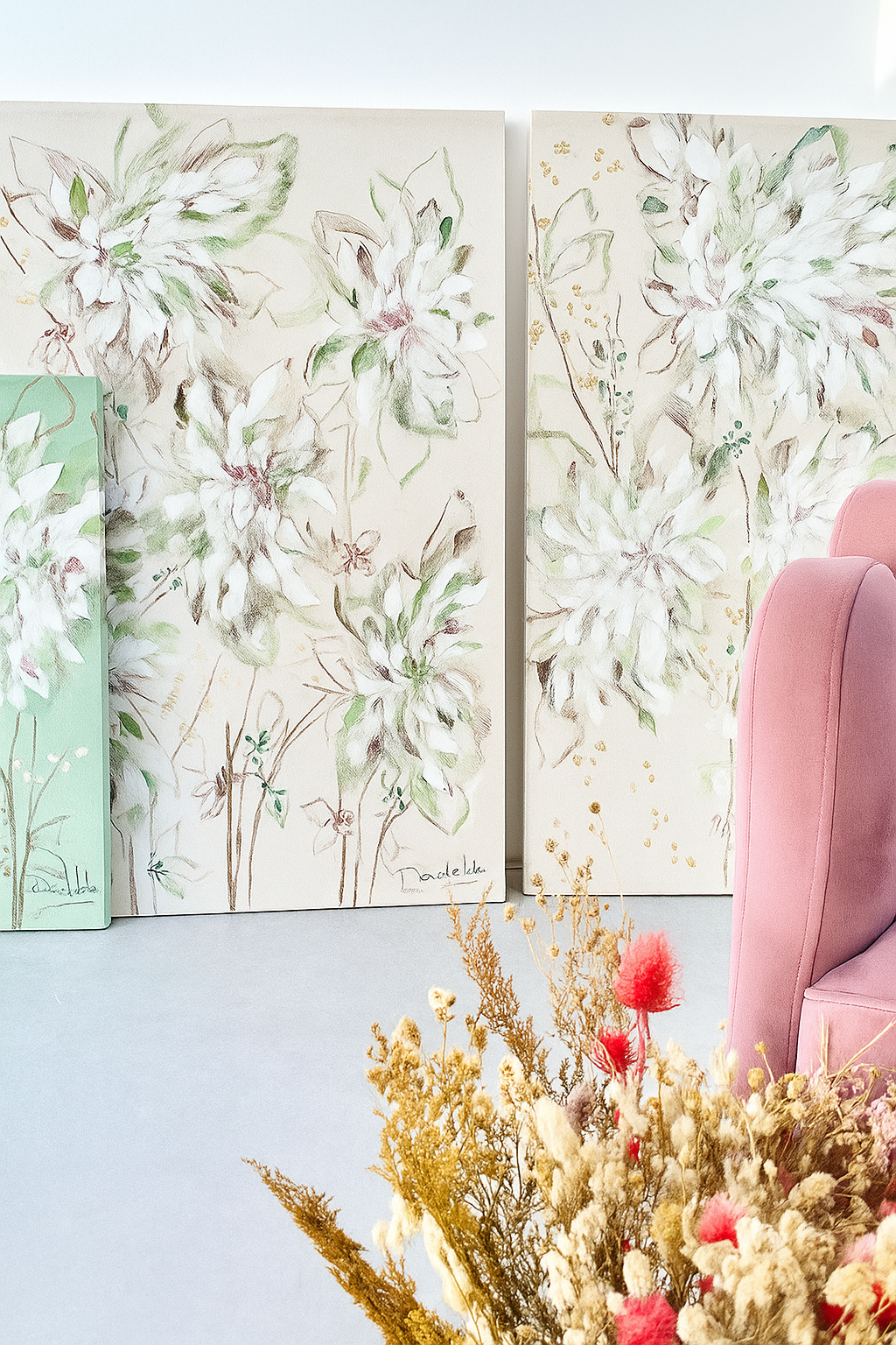 Dos cuadros de estilo tropical con hojas verdes y detalles rosados apoyados en la pared, al lado de un sillón rosa aterciopelado.