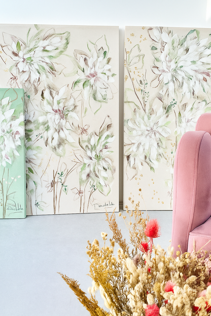 Dos cuadros de estilo tropical con hojas verdes y detalles rosados apoyados en la pared, al lado de un sillón rosa aterciopelado.