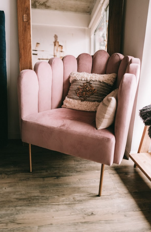 Sillón tapizado en terciopelo rosa con un diseño de respaldo en forma de concha, formado por paneles verticales acolchonados. Tiene patas delgadas metálicas doradas y está decorado con dos cojines: uno beige y otro con textura y diseño bordado en tonos tierra