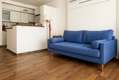Sillón de tres cuerpos en tapizado azul, con apoyabrazos rectos, patas de madera y almohadones cilíndricos a los costados, ubicado en un living moderno con piso de madera.