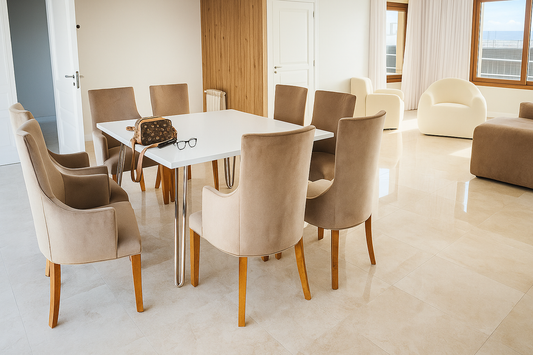 Comedor moderno con mesa rectangular blanca de patas metálicas cromadas y ocho sillas tapizadas en beige claro, sobre piso de mármol pulido y con gran entrada de luz natural.