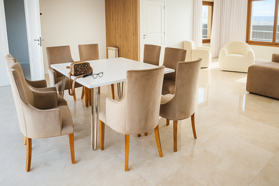 Comedor moderno con mesa rectangular blanca de patas metálicas cromadas y ocho sillas tapizadas en beige claro, sobre piso de mármol pulido y con gran entrada de luz natural.