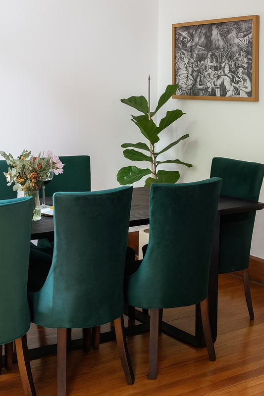 Comedor elegante con mesa rectangular de madera oscura y seis sillas de terciopelo verde esmeralda, acompañado de un florero central con flores frescas e iluminado por luz natural.