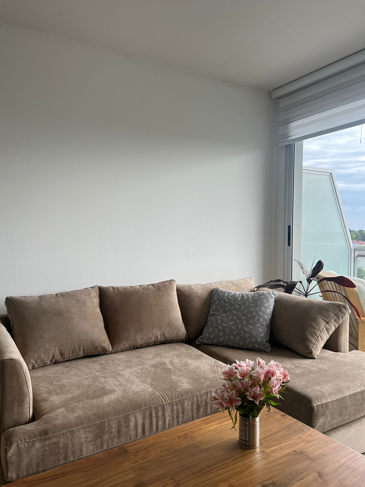 Sofá esquinero en tono beige con cojines a juego, acompañado por un almohadón gris estampado. Frente a él se ubica una mesa de madera con un florero con flores rosadas, en un ambiente luminoso con ventanal.