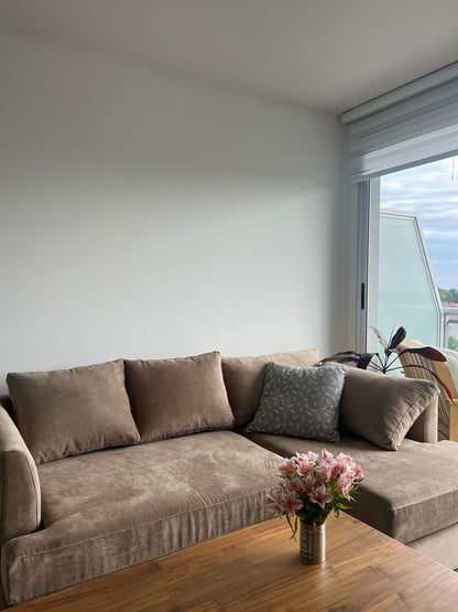 Sofá esquinero en tono beige con cojines a juego, acompañado por un almohadón gris estampado. Frente a él se ubica una mesa de madera con un florero con flores rosadas, en un ambiente luminoso con ventanal.