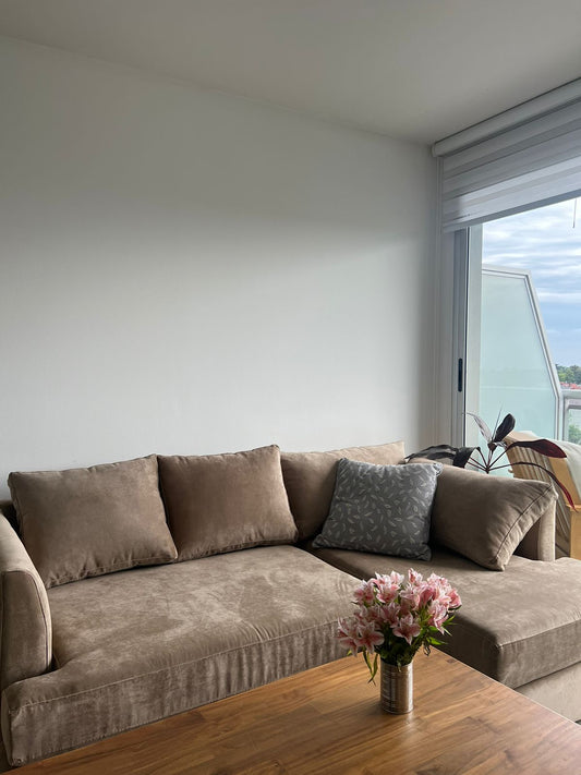 Sofá esquinero en tono beige con cojines a juego, acompañado por un almohadón gris estampado. Frente a él se ubica una mesa de madera con un florero con flores rosadas, en un ambiente luminoso con ventanal.