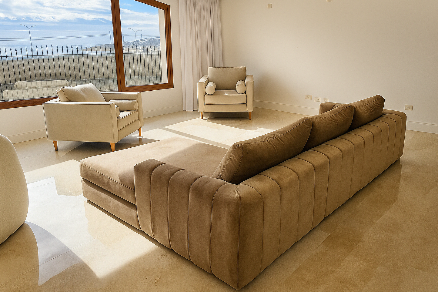 Living luminoso con sillón modular marrón aterciopelado y dos butacas tapizadas en tono beige, frente a un gran ventanal con vista al exterior.