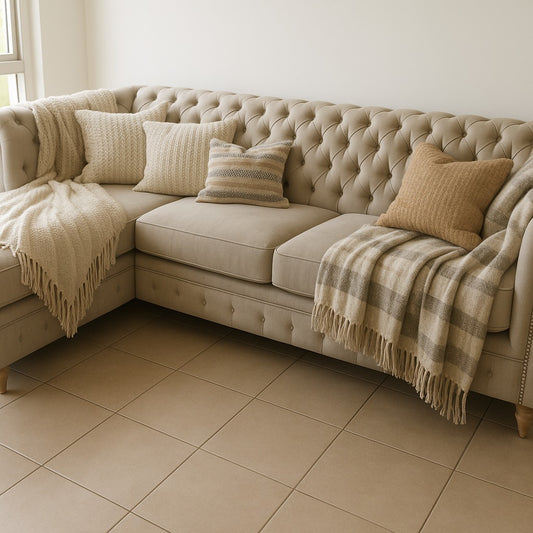 fá esquinero estilo Chesterfield tapizado en tela beige claro, con diseño capitoné en el respaldo y brazos enrollados. El sofá tiene patas de madera torneadas en tono natural, y está decorado con cojines en tonos tierra y mantas tejidas en colores neutros, que cuelgan suavemente sobre los brazos.